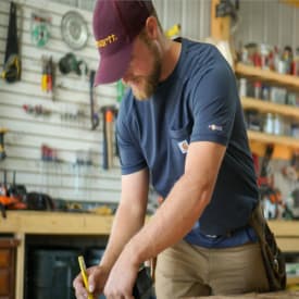 Carhartt: A man wearing a maroon Carhartt baseball cap and a Carhartt Dark Cobalt Blue Heather K87 pocket t-shirt does a measuring task at a workbench. 