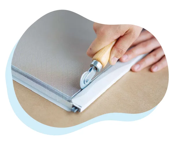 A close-up of a person's hands as they cut along the edge of a screen for screen repair and fabrication.