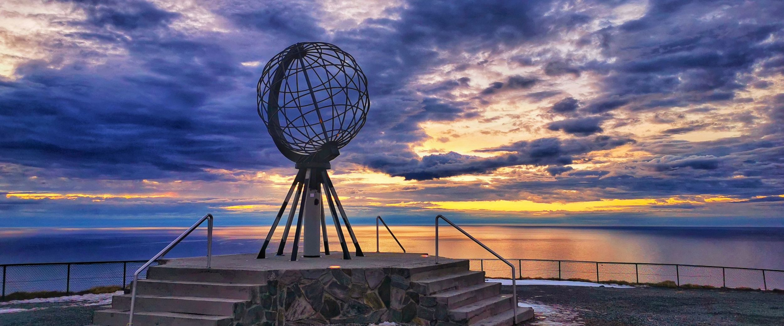 The Globe monument at the North Cape standing on a cliff above the sea, with dramatic clouds and colourful sunset light on the horizon.