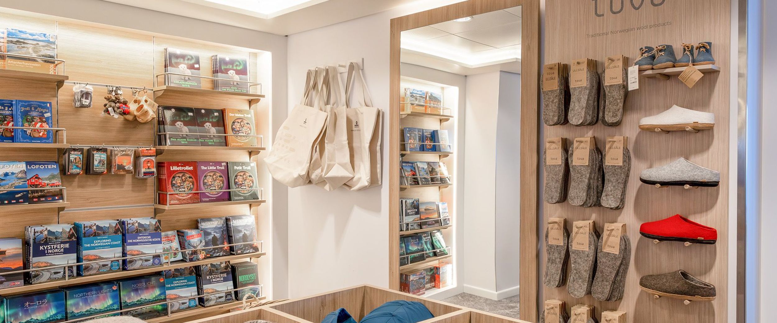 Interior of Havila Shop showing shelves with books and local products, canvas tote bags, folded blankets in a wooden display table, and a wall of warm slippers and wool socks.