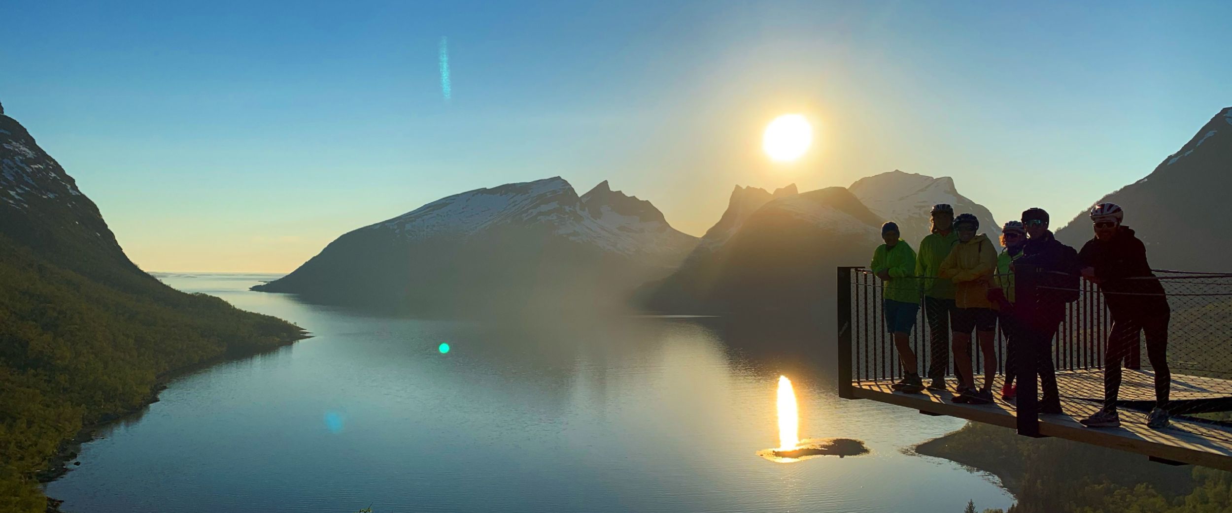 Golden light from the low Arctic sun reflects across a calm fjord on Senja, Norway, as a small group pauses at a mountain viewpoint to take in the vast landscape. Snow-dusted peaks, deep blue water, and fresh spring greenery come together in a quiet moment where nature completely steals the show.