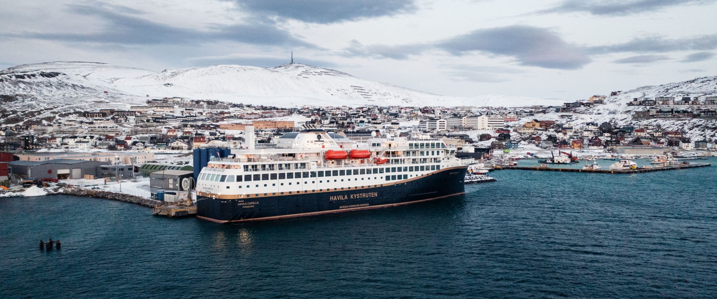 The Havila Voyages ship Havila Capella is docked in a snow-covered coastal town, with wintery mountains in the background and cold blue sea in the foreground.