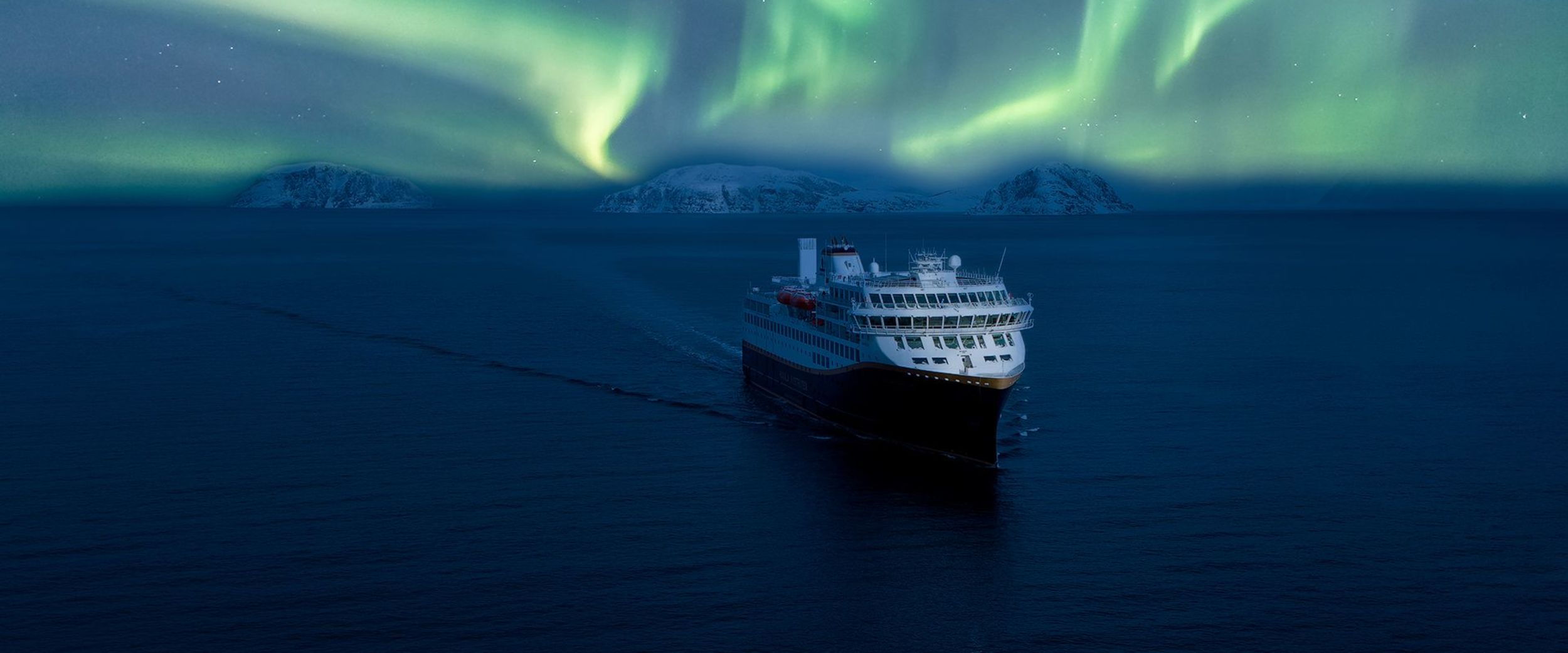 A Havila Voyages ship sails silently through Arctic waters beneath a vivid display of the Northern Lights, surrounded by snow-covered mountain silhouettes along Norway’s northern coast.