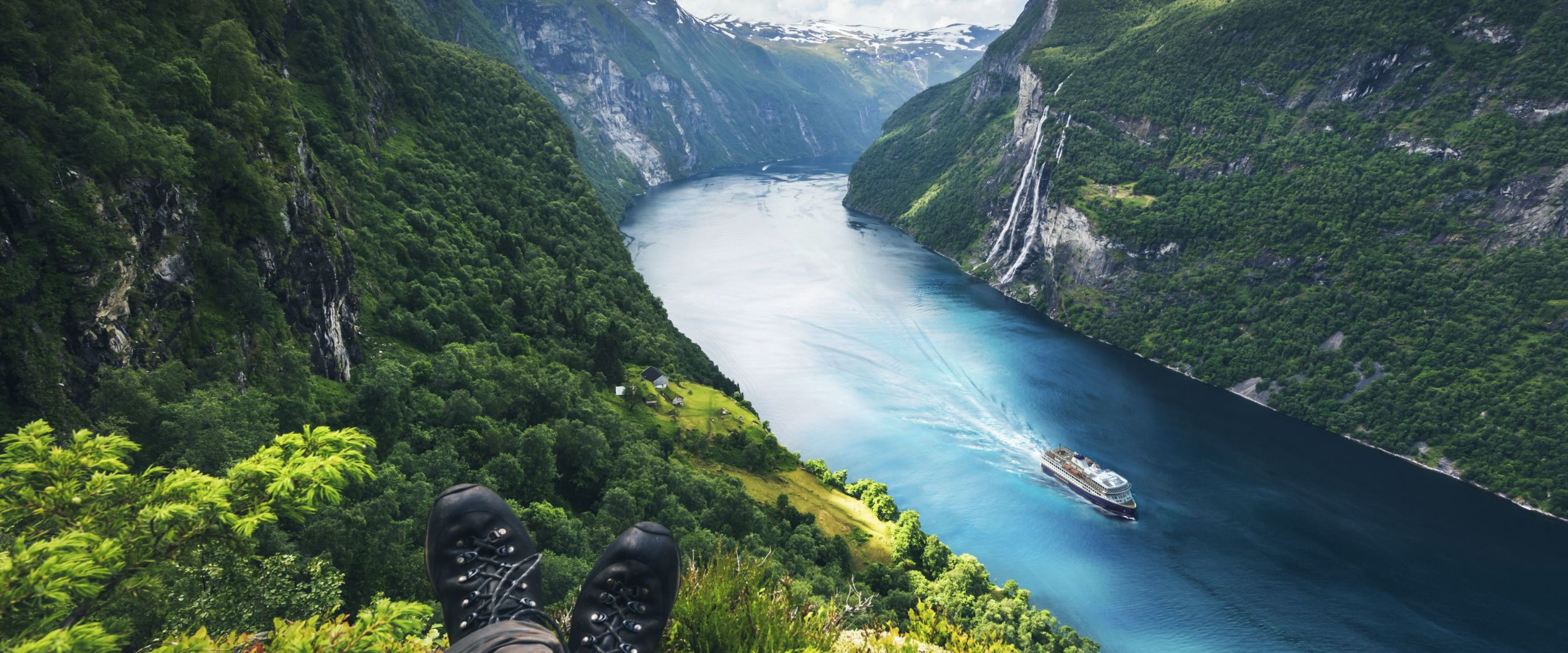 Havila ship sailing in the azure blue Geiranger fjord with steep green sides and a partly cloudy sky.