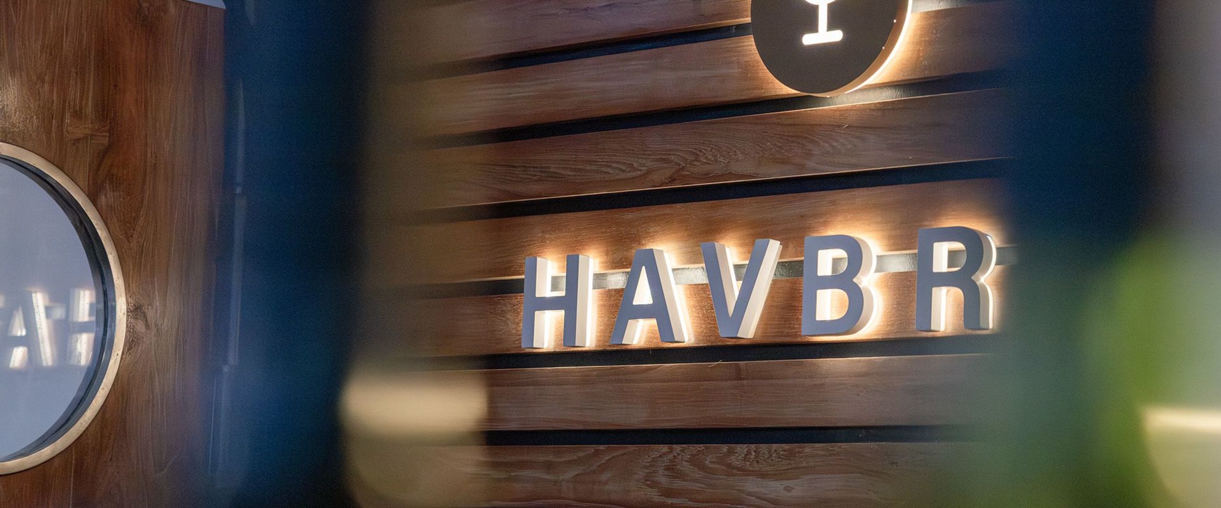 Soft-focus view of the illuminated "Havbris Bar" bar sign on a wooden wall aboard a Havila Voyages ship, creating a warm and atmospheric interior scene.