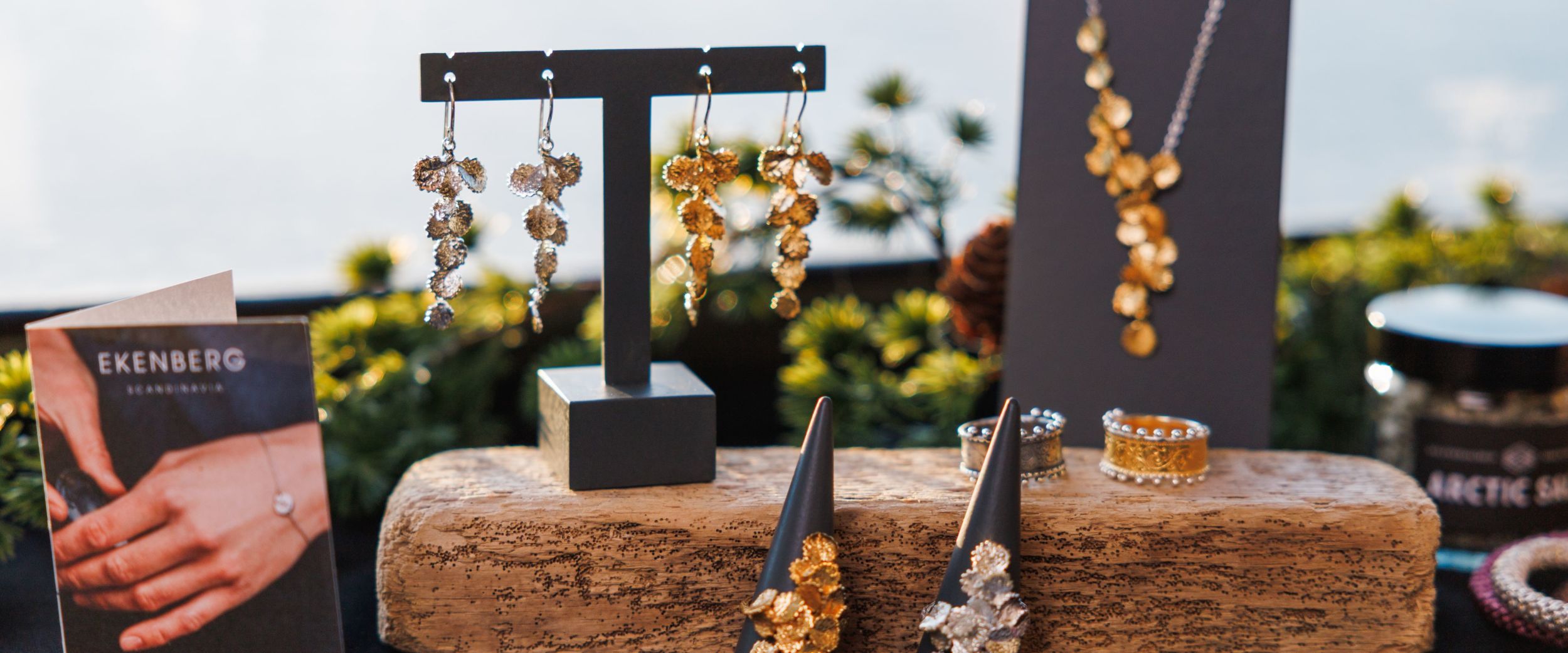 Display of gold and silver Ekenberg “Fjords of Norway” jewellery on a rustic wooden block, including dangling earrings, a pendant necklace and rings, with greenery and the sea softly blurred in the background.