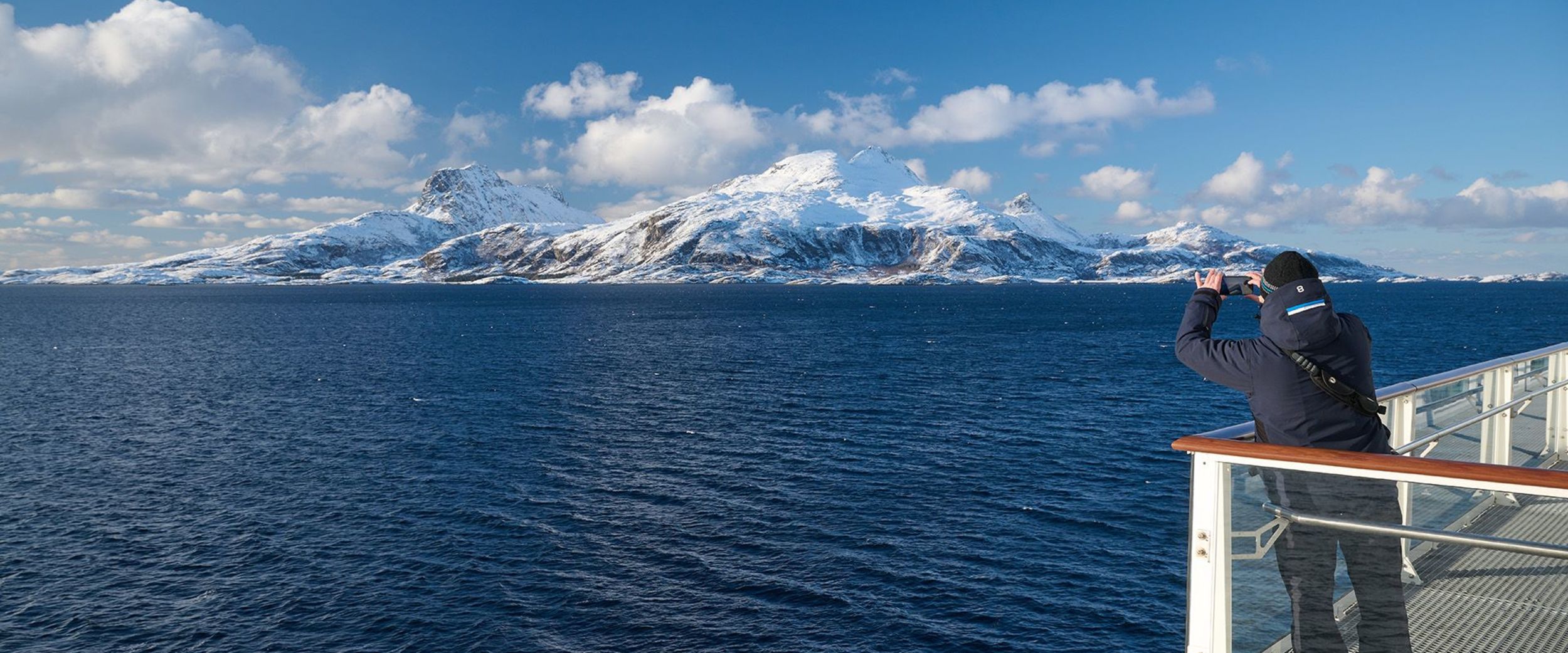 A passenger photographing a snow-covered mountain landscape from the deck of a Havila Voyages ship on a clear, sunny day.