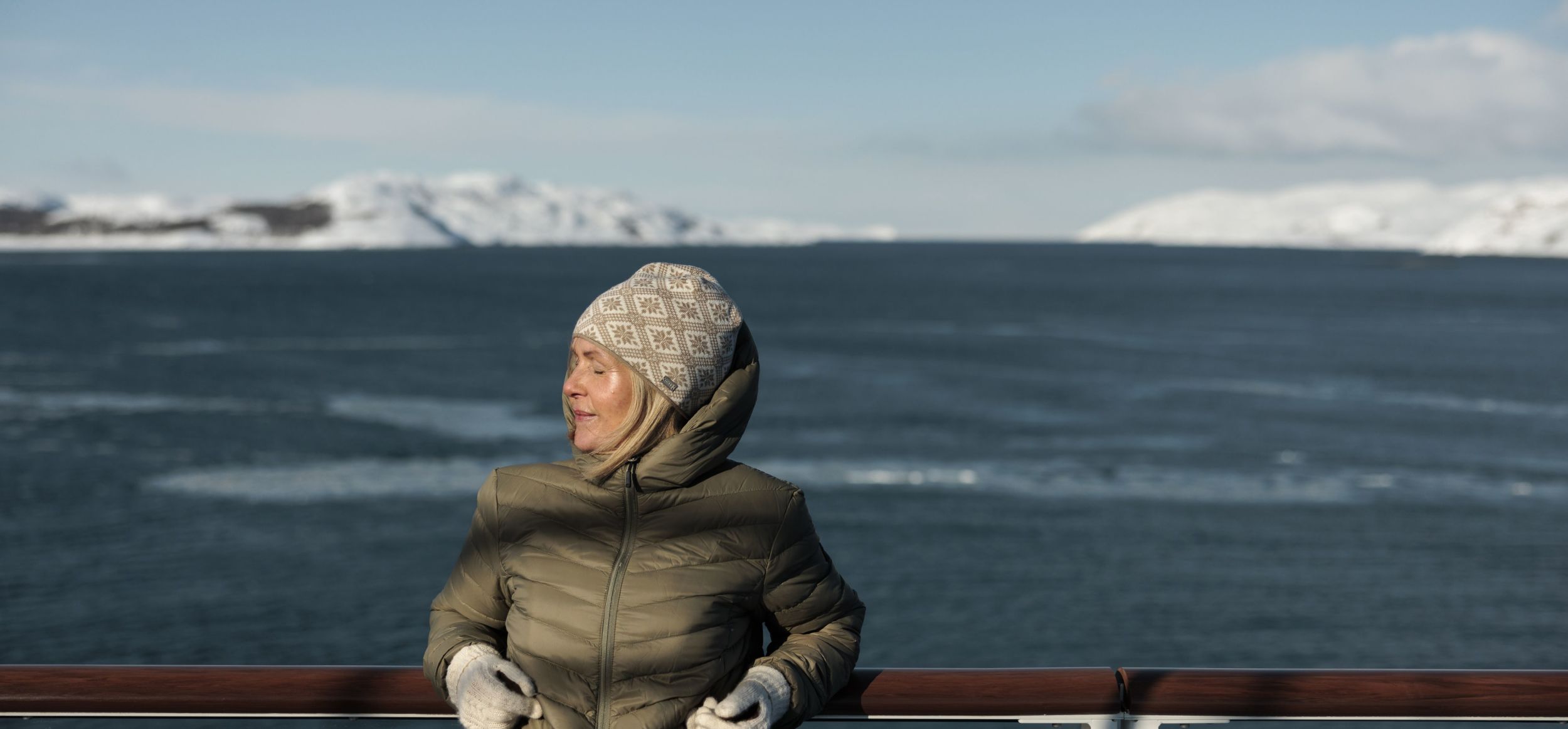 Woman in winter clothing enjoying the view from a cruise ship deck, with snowy mountains and a calm Norwegian fjord in the background.