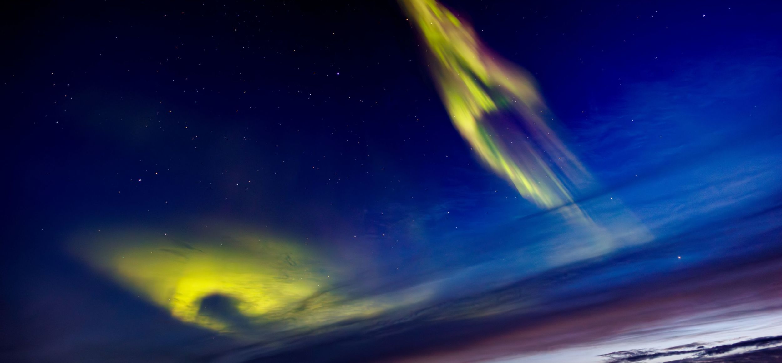 Northern Lights in vivid green and yellow dance across a starry night sky over dark mountain silhouettes, with streaks of colour reflecting the Arctic atmosphere.