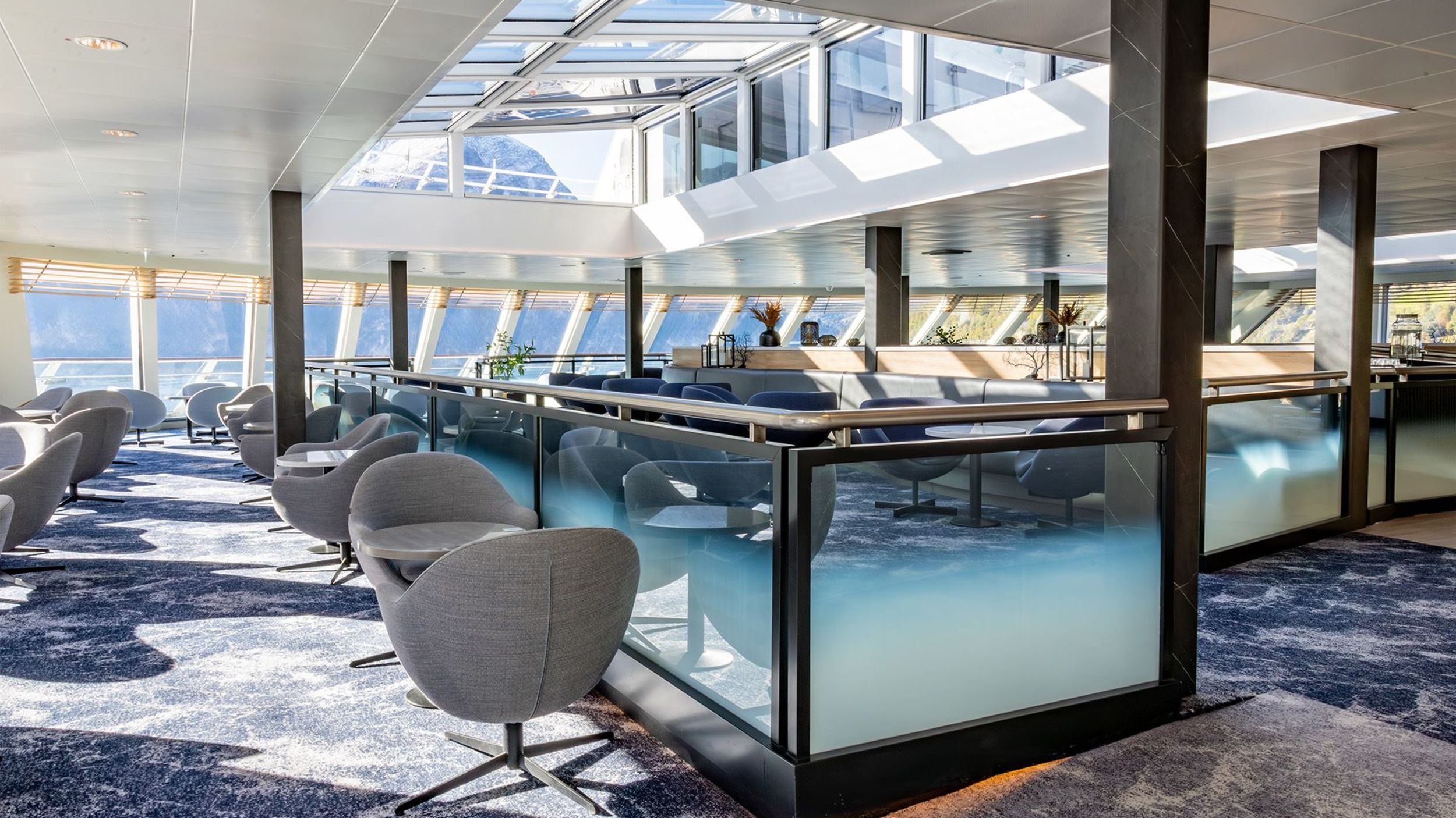 Interior of Havblikk Bar & Lounge on deck nine of a Havila Voyages ship, featuring modern lounge chairs, floor-to-ceiling windows, and a glass roof allowing natural light to flood the space with mountain views in the background.