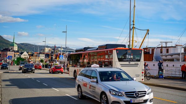 Cars and a city bus driving along a street in Bergen near the harbour, with boats, buildings and mountains in the background under a blue sky.