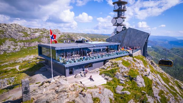 Modern mountaintop restaurant with outdoor terrace on Mount Ulriken in Bergen, Norway. A cable car is arriving, and the TV tower rises behind the building, with dramatic mountain scenery in the background.