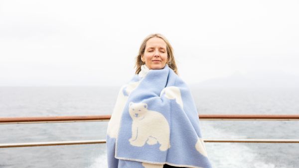 Woman standing on the open deck of a Havila Voyages ship, eyes closed and smiling, wrapped in a light blue blanket with white polar bear pattern, with the sea and misty coastline in the background.