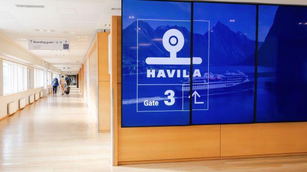 Digital screen showing Havila Voyages Gate 3 in a ferry terminal corridor, with passengers walking toward the boarding gates.