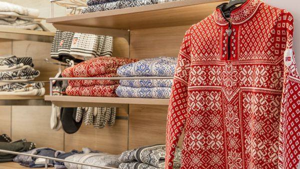 Display of patterned Norwegian wool sweaters in Havila Shop, with a red traditional knit sweater featuring a small Norwegian flag patch hanging in the foreground.