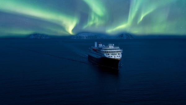 A Havila Voyages ship sails silently through Arctic waters beneath a vivid display of the Northern Lights, surrounded by snow-covered mountain silhouettes along Norway’s northern coast.