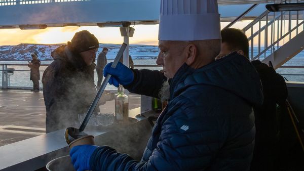 Chef in gear serving hot soup from a steaming pot on the outer deck during sunset, with snowy mountains and sea in the background.
