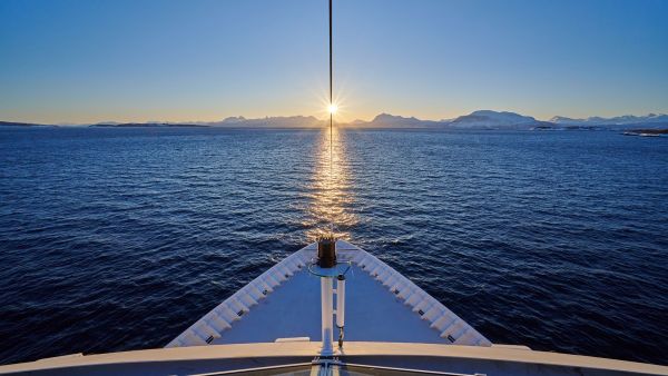 View from the bow of a Havila Voyages ship sailing toward the horizon at sunset, with the sun perfectly aligned over the sea and snowy mountains in the distance.