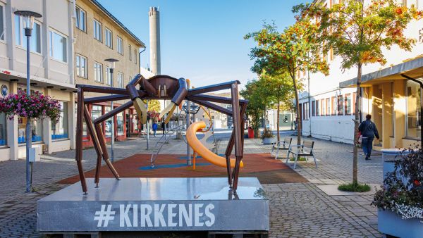 A large king crab sculpture in the centre of Kirkenes, set along a quiet pedestrian street with buildings, trees, and a small playground in the background.