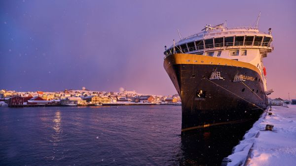 Havila coastal cruise ship docked in a snowy Norwegian harbor at sunset, with warm pastel skies and a coastal town in the background.
