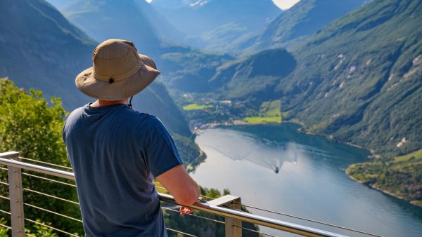 Traveler wearing a sun hat at a viewpoint overlooking a fjord and mountains.