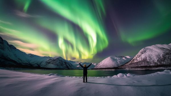 A lone figure stands on a snowy shore, arms outstretched, as vivid green auroras dance across the night sky above mountains and a tranquil fjord.