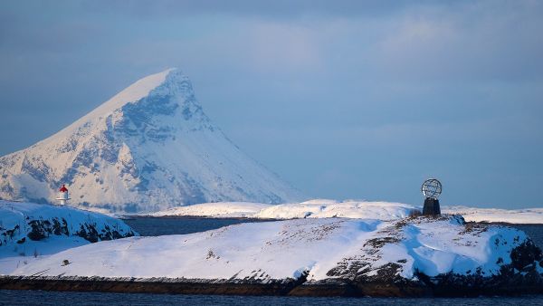 Snow-covered coastal island in Norway featuring the Arctic Circle Globe monument, with a dramatic mountain peak in the background under a cloudy sky.