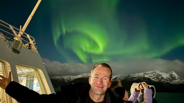 Smiling passenger on the deck of a ship under bright green northern lights, with snow-covered mountains and another traveler taking a photo in the background.