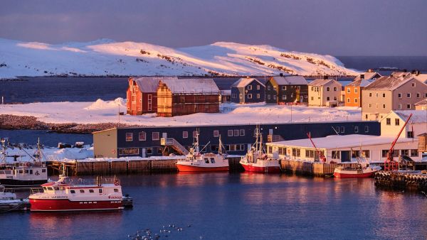 Colorful fishing boats and snow-covered buildings in the harbor of a Norwegian coastal town, bathed in soft snowy light.