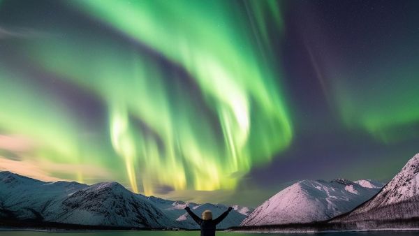 A person stands on snow-covered ground with arms raised under a spectacular northern lights display, surrounded by snow-covered mountains and a calm fjord.