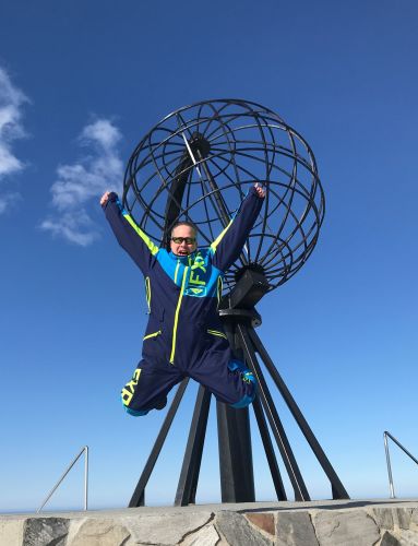 Guest in a blue overall jumping with arms raised in front of the Globe monument at the North Cape on a clear, sunny day.