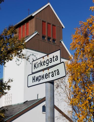 Street sign reading “Kirkegata” and “Киркегата” in front of Kirkenes Church, surrounded by golden autumn trees under a clear blue sky.