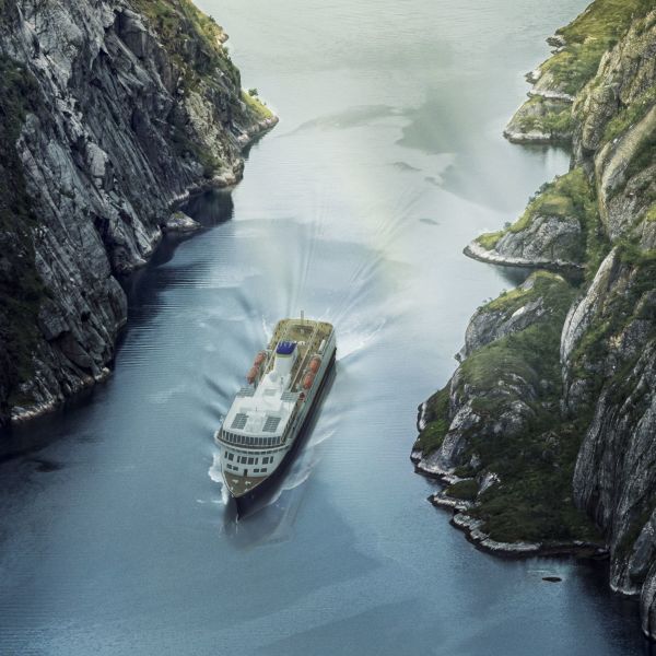 Havila ship sailing through narrow fjord with steep stony sides and some green vegetation. 