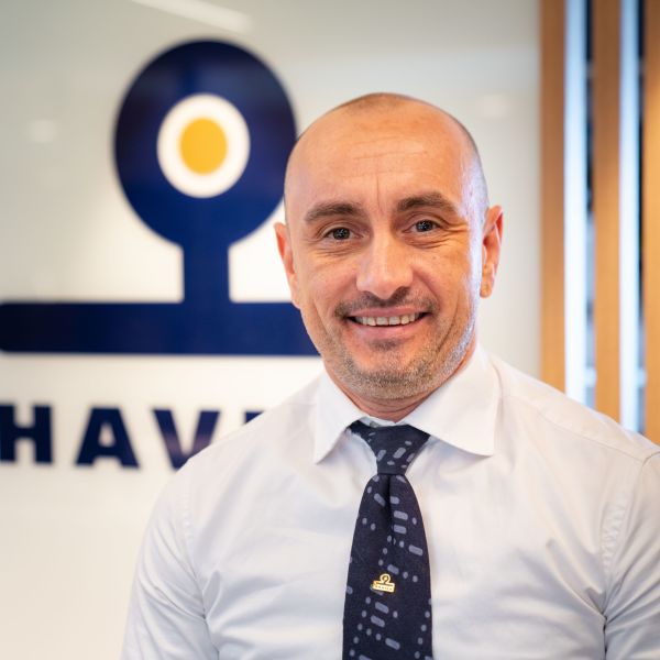 A smiling man wearing a white shirt and a dark blue tie stands in front of a glass wall featuring the Havila Voyages logo. He is bald with light stubble.  The surrounding environment is bright and modern with wooden details.