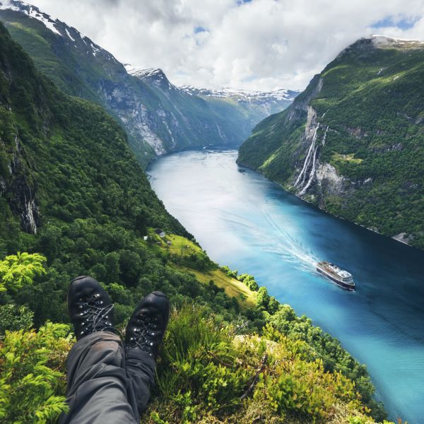 Havila ship sailing in the azure blue Geiranger fjord with steep green sides and a partly cloudy sky.