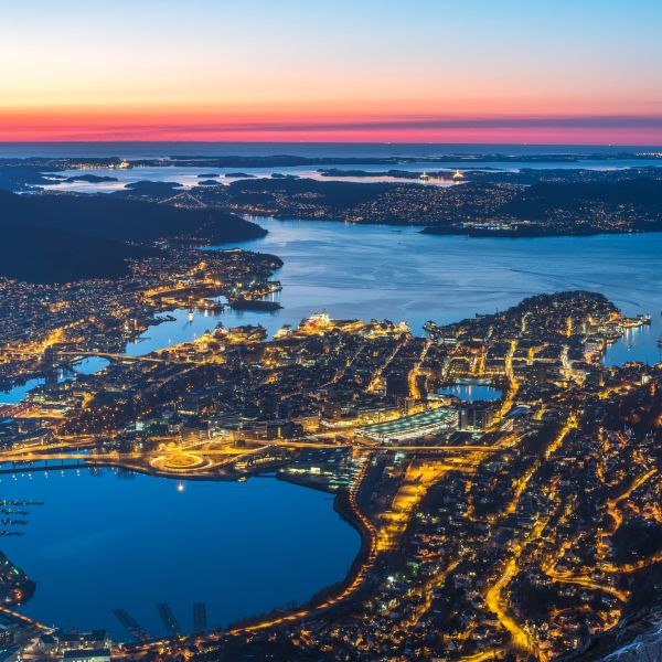 Evening view over Bergen, Norway, captured from above. The city is illuminated with warm lights, reflecting in the fjords, with a colorful sunset fading into twilight over the mountains and coastal islands.