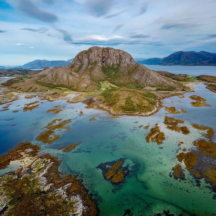 Drone photo of Torghatten near Brønnøysund, surrounded by turquoise shallow waters, skerries, and small islands under a cloudy sky.