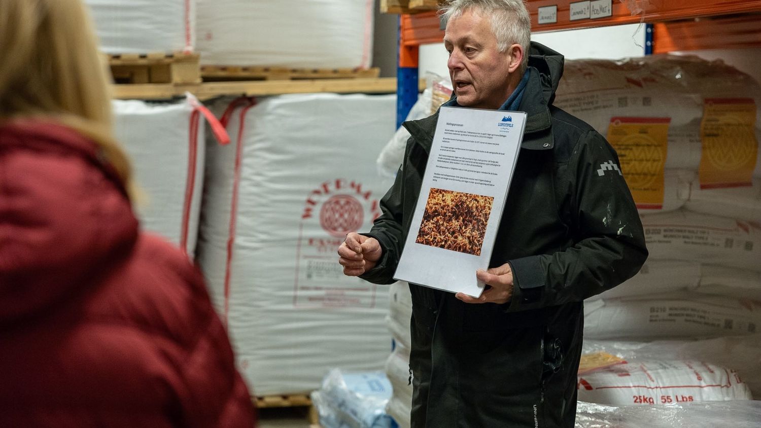A guide at Lofotpils brewery explaining the beer production process to visitors, holding an information sheet in a warehouse filled with large sacks of ingredients.