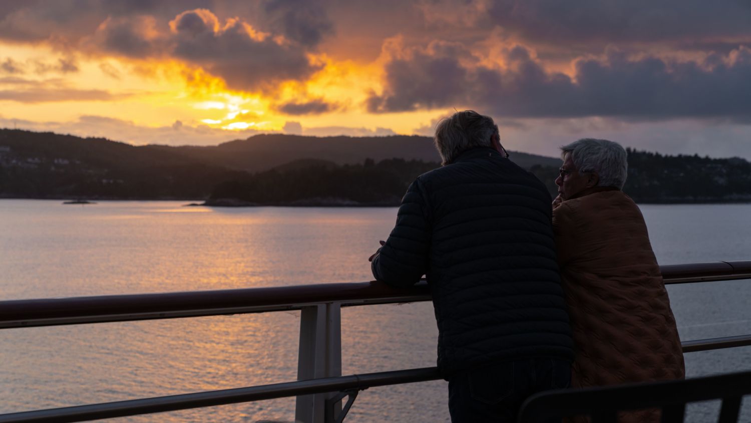 A couple stands close together on the deck of a ship, watching a golden sunset over calm waters. The man is leaning on the railing, while the woman is wrapped in a warm brown jacket, leaning toward him. In the background, low mountains and dramatic clouds are illuminated by the fading light.