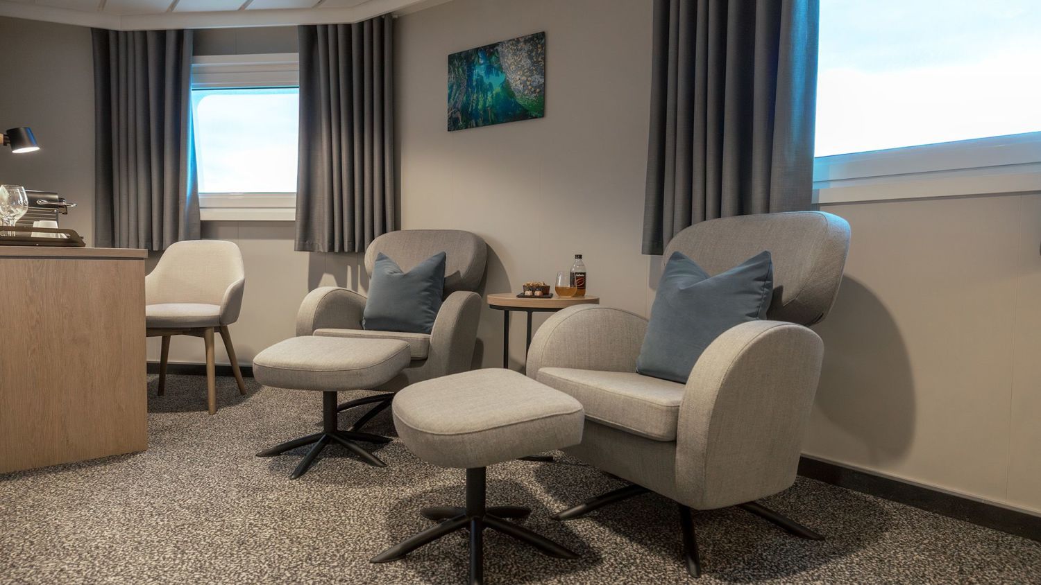 Modern mini suite on board a ship, featuring two comfortable armchairs with footstools, a small side table with drinks and snacks, and large windows offering a sea view. The interior is decorated in neutral tones with grey curtains, wall art, and soft lighting, creating a cozy and upscale atmosphere.