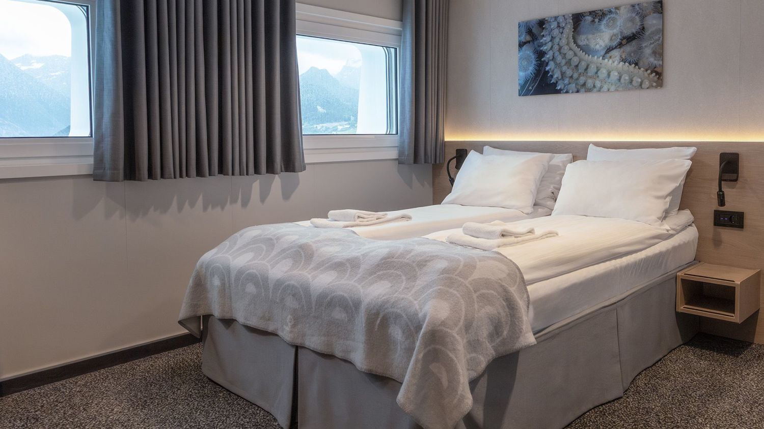 Elegant double cabin with mountain views through two large windows, featuring a soft bed with white linens, grey curtains, warm lighting, and coastal-inspired artwork above the headboard.