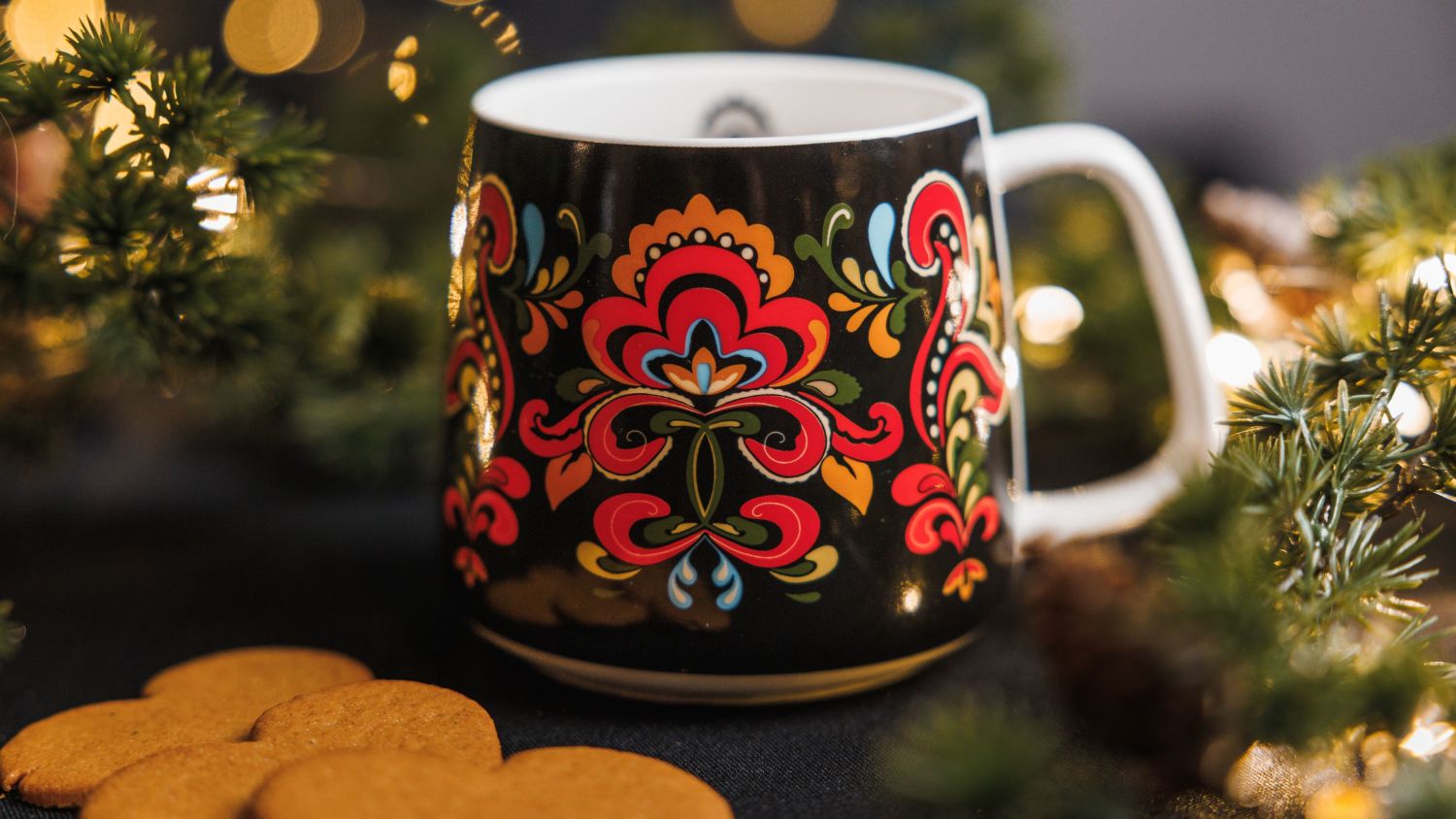 Decorative black mug with a colorful Norwegian folk pattern placed beside gingerbread cookies and evergreen branches, with warm blurred Christmas lights in the background.