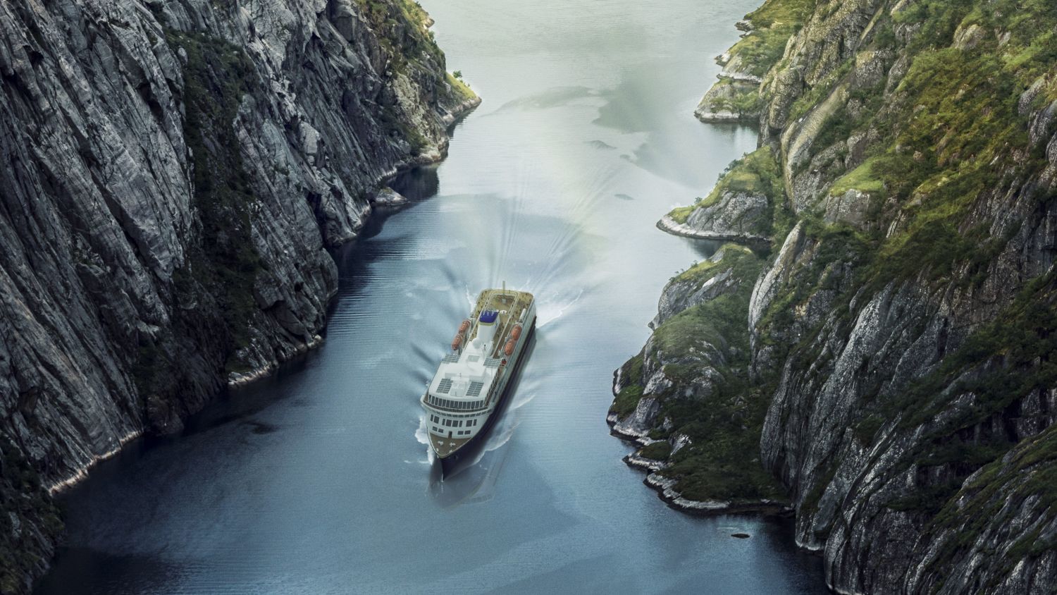 Havila ship sailing through narrow fjord with steep stony sides and some green vegetation. 