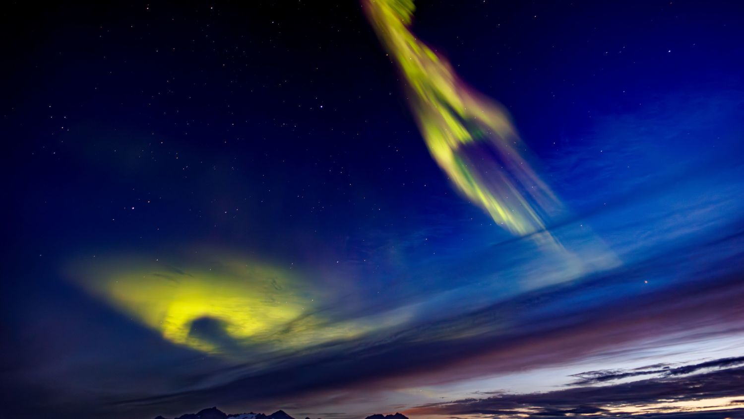 Northern Lights in vivid green and yellow dance across a starry night sky over dark mountain silhouettes, with streaks of colour reflecting the Arctic atmosphere.