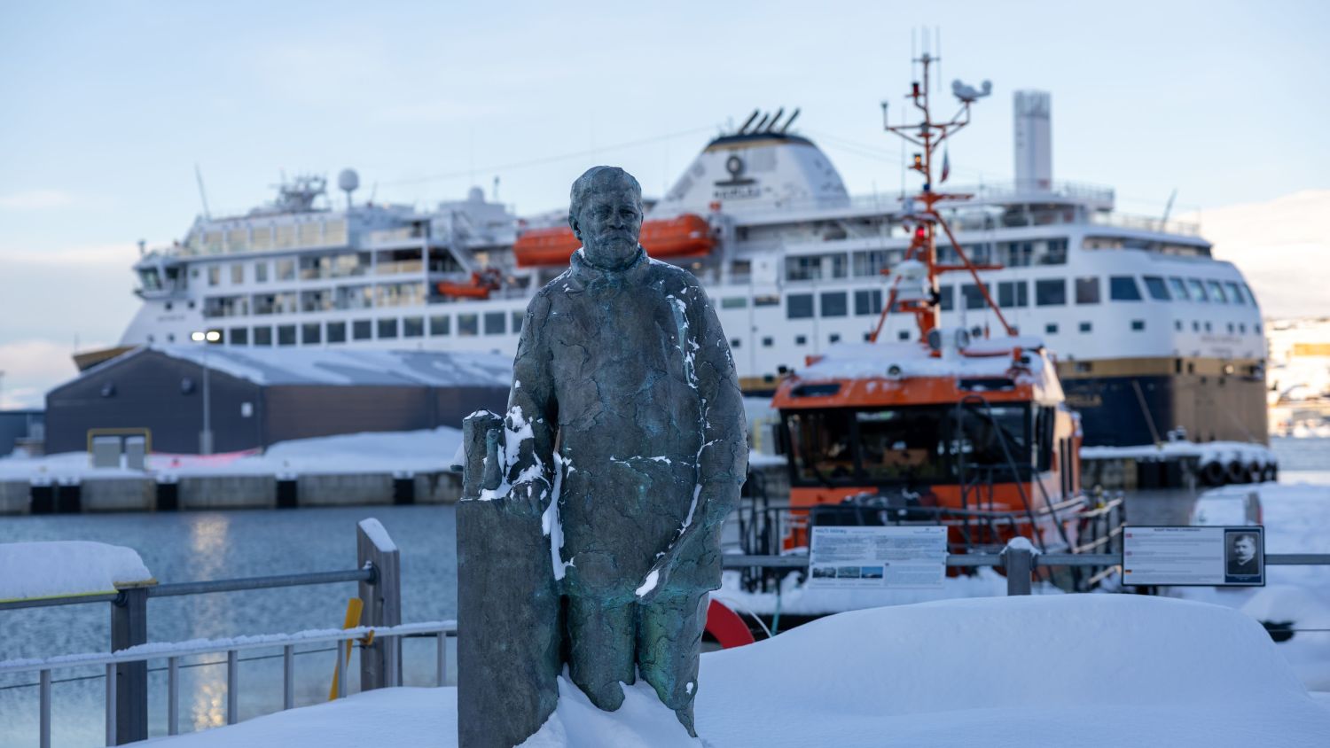 A statue covered in snow stands in the foreground at Hammerfest harbor, with a Havila Voyages ship docked in the background alongside smaller vessels and port infrastructure.