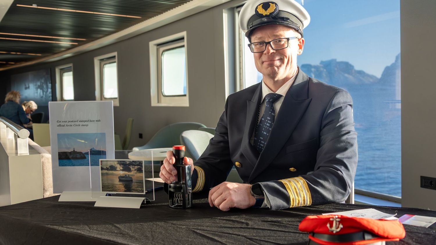 Crew member sitting at a table with postcards onboard a Havila ship, with views of the Norwegian coast through the window.