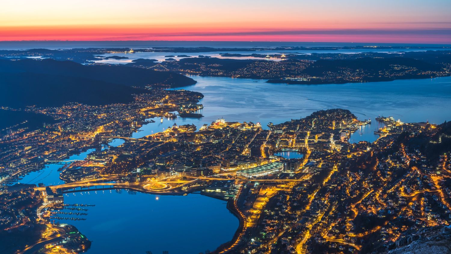 Evening view over Bergen, Norway, captured from above. The city is illuminated with warm lights, reflecting in the fjords, with a colorful sunset fading into twilight over the mountains and coastal islands.