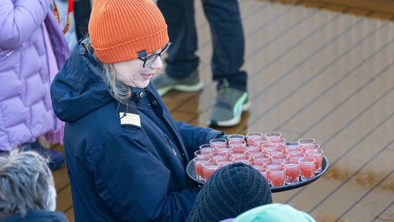 Crew member serving small glasses of a red drink to guests on deck during the Arctic Circle crossing ceremony onboard a Havila ship.