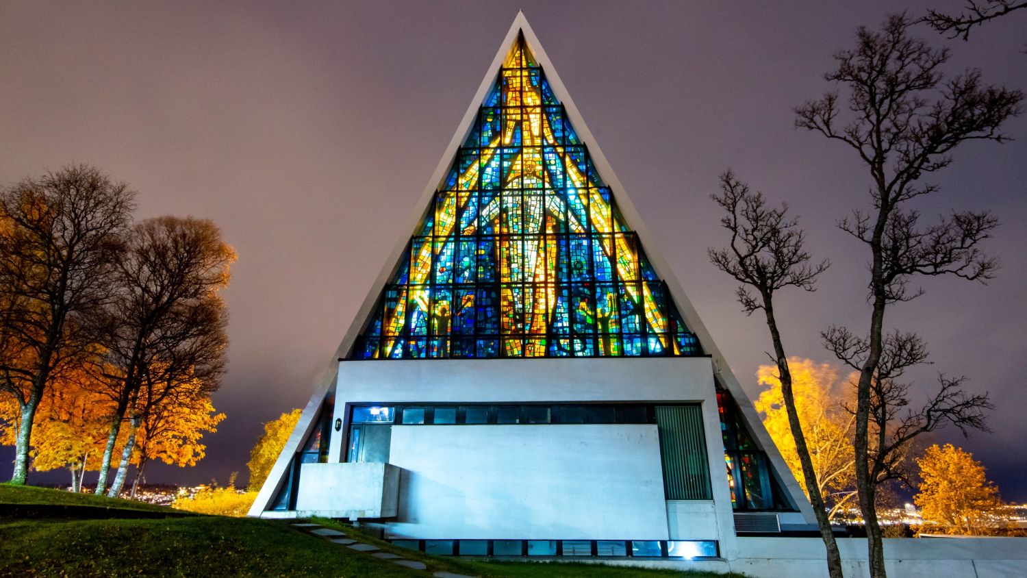 Arctic Cathedral in Tromsø illuminated on an autumn evening, with colorful stained-glass windows glowing through the triangular façade