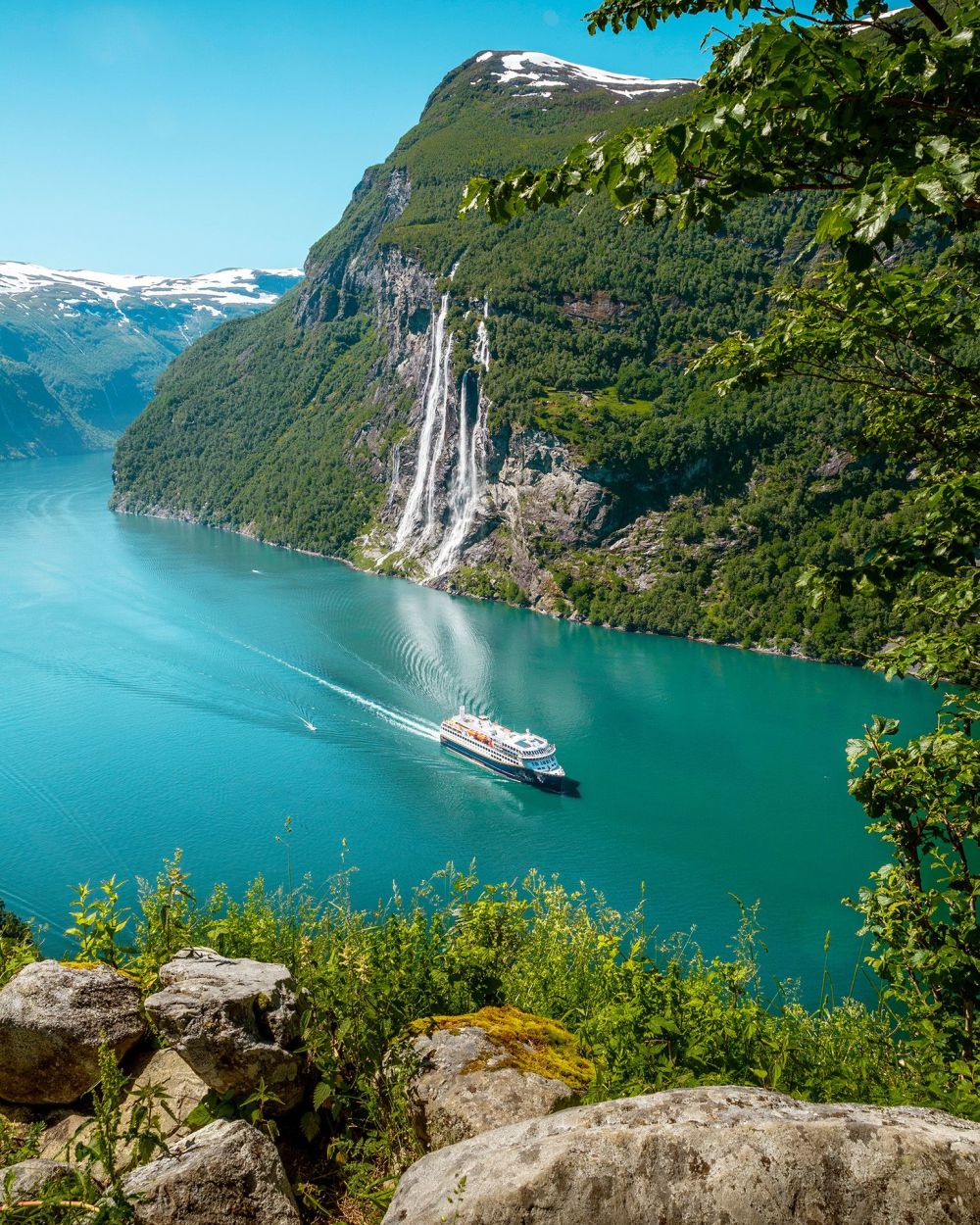 Havila Voyages ship sailing through a turquoise fjord beneath steep green mountains and waterfalls.